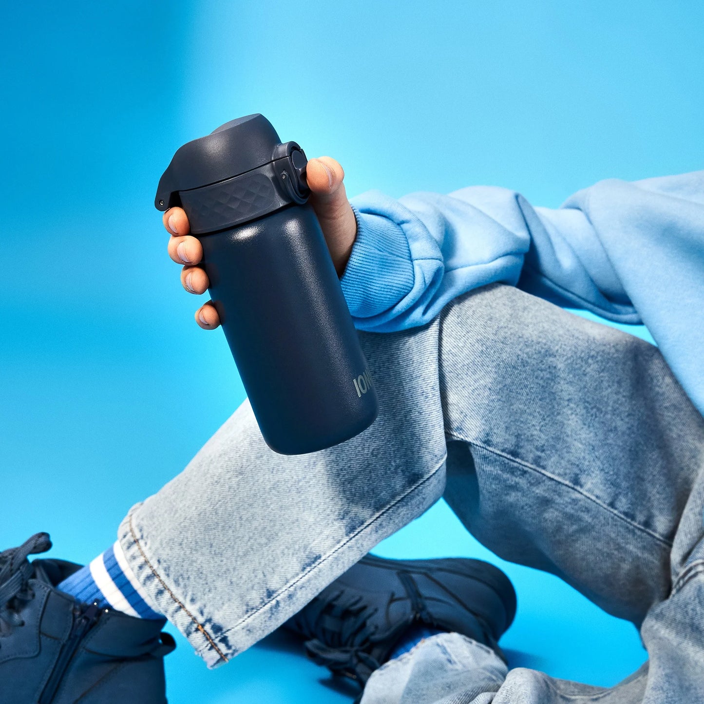 A hand holds a dark blue bottle labeled "Ion8" against a blue background worn by a person in blue jeans and a light blue sweatshirt sitting cross-legged on the floor.