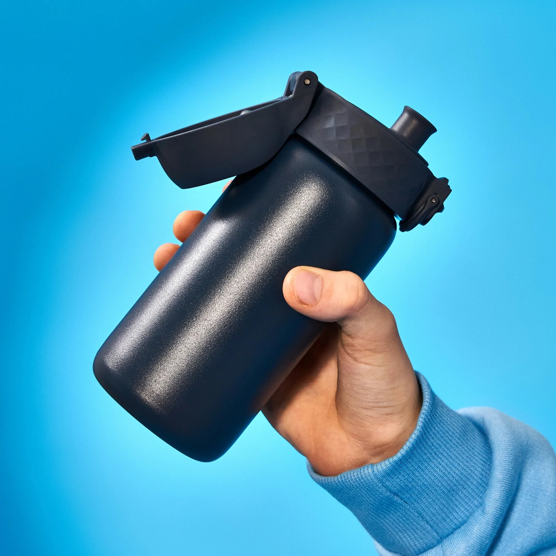 A black bottle with a flip-top lid is held in a hand wearing a blue sleeve set against a bright blue background.