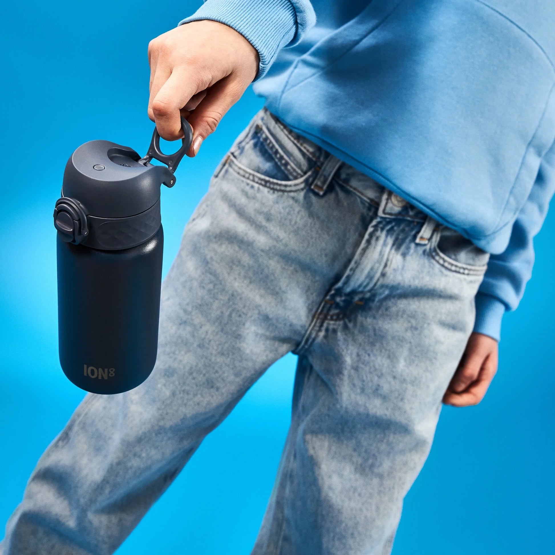 A navy blue water bottle is held by a hand against a bright blue background. The person wearing light blue jeans and a blue sweatshirt. Text: "ION8"