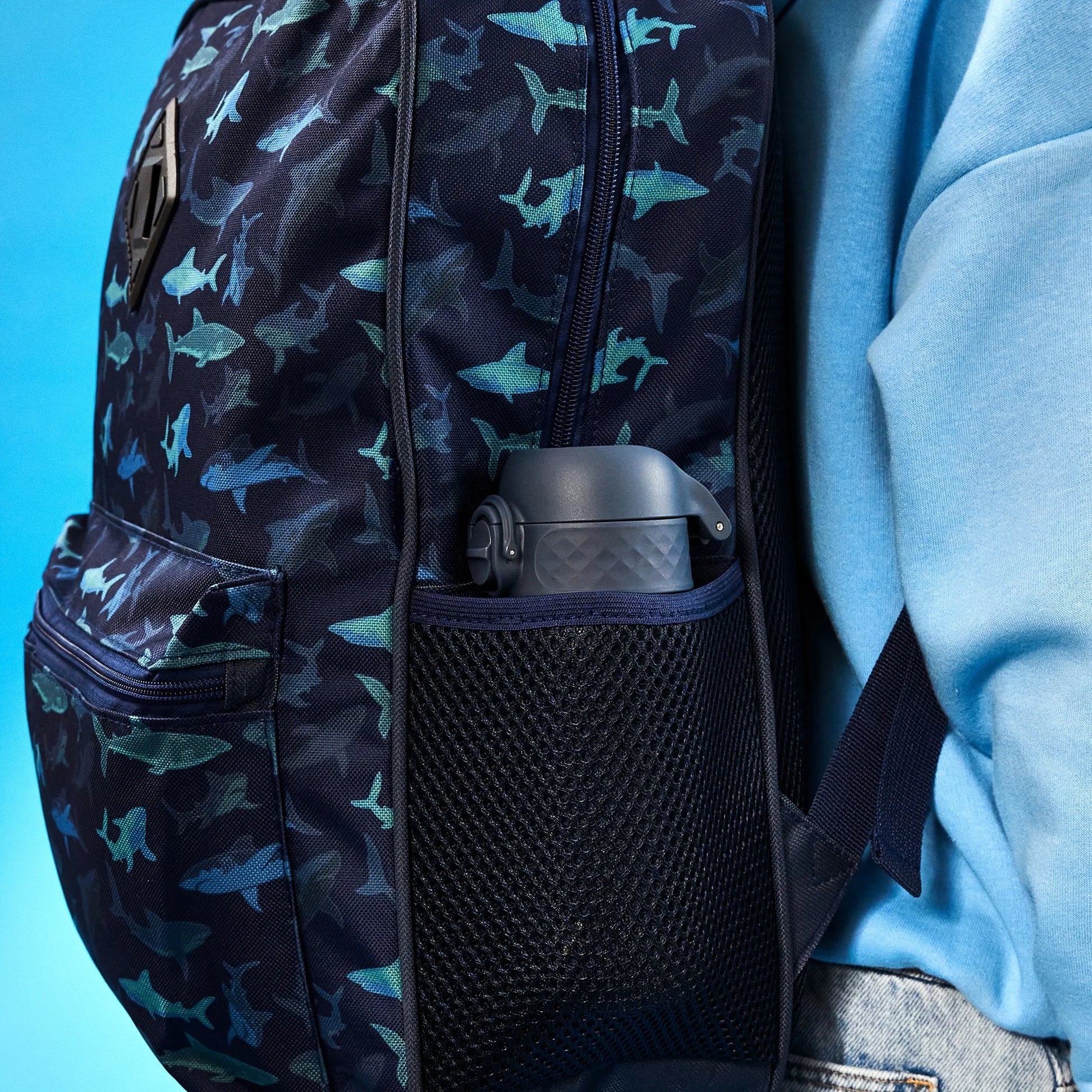 A colorful backpack with shark patterns holds a dark water bottle in its mesh side pocket against a person wearing a blue shirt in front of a blue background.