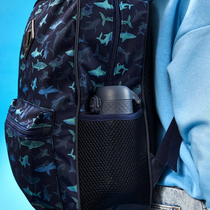 A colorful backpack with shark patterns holds a dark water bottle in its mesh side pocket against a person wearing a blue shirt in front of a blue background.