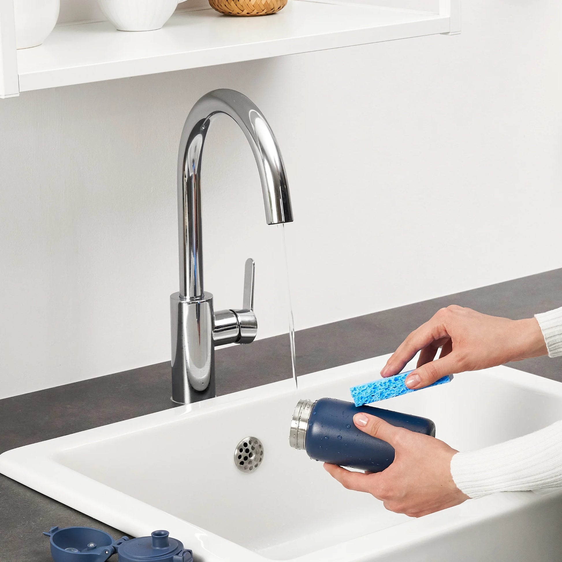 Hands cleaning a blue bottle with a sponge under running water from a chrome kitchen faucet in a white sink surrounded by minimalistic kitchen decor.