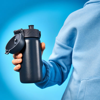 A hand holds a black water bottle with an open lid against a bright blue background wearing a light blue hoodie.