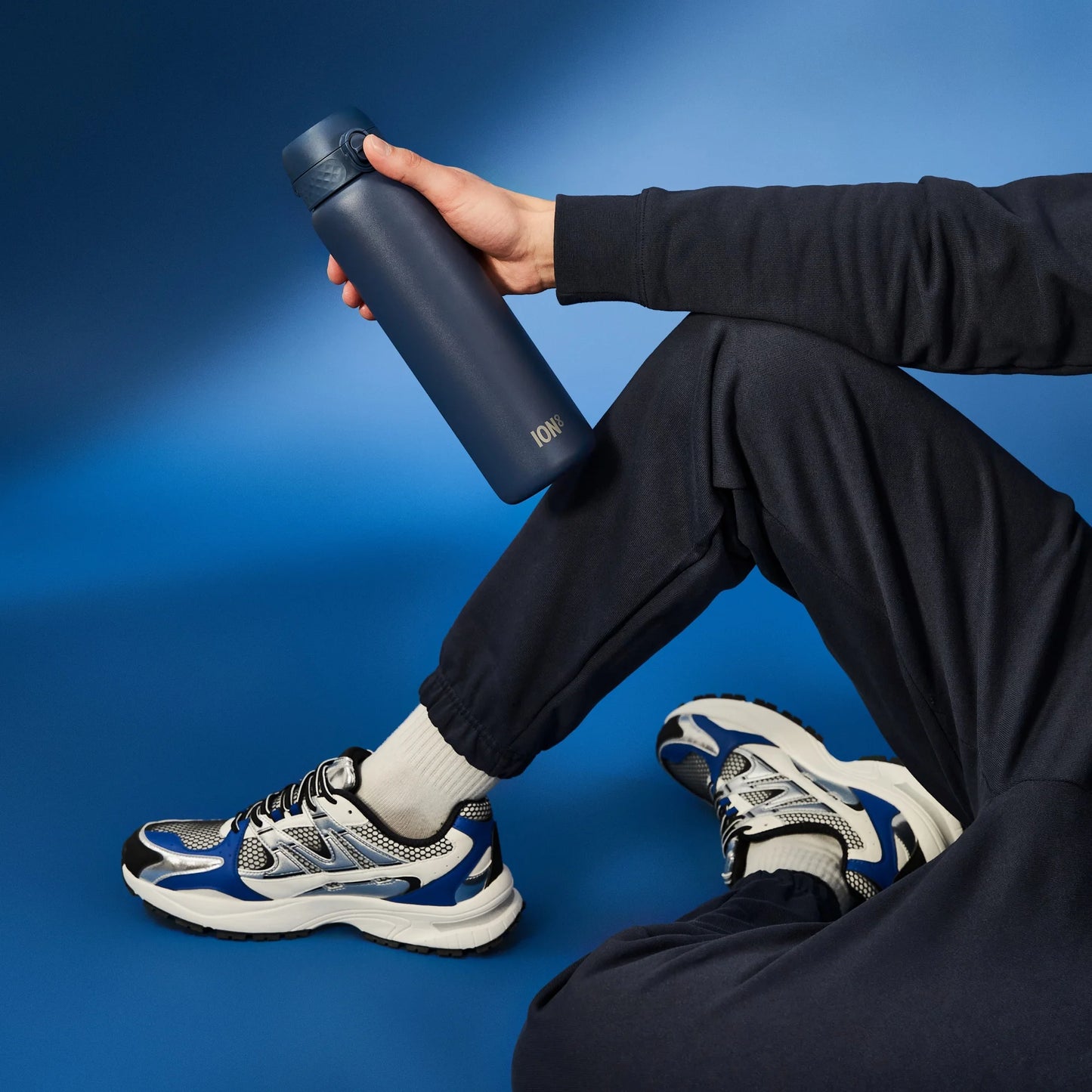 A person holds a dark blue water bottle labeled "ION8" while seated on a blue floor wearing black pants and blue-and-white sneakers.