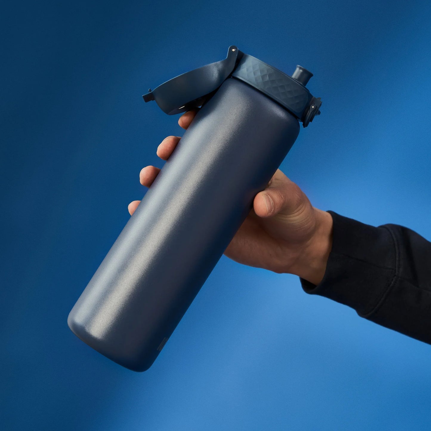 Hand holding a blue insulated water bottle with a flip-top lid against a solid blue background.