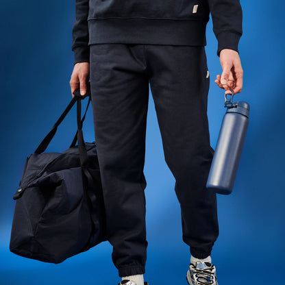 Person holding a navy gym bag and a blue water bottle wearing a black tracksuit and sneakers against a solid blue background.