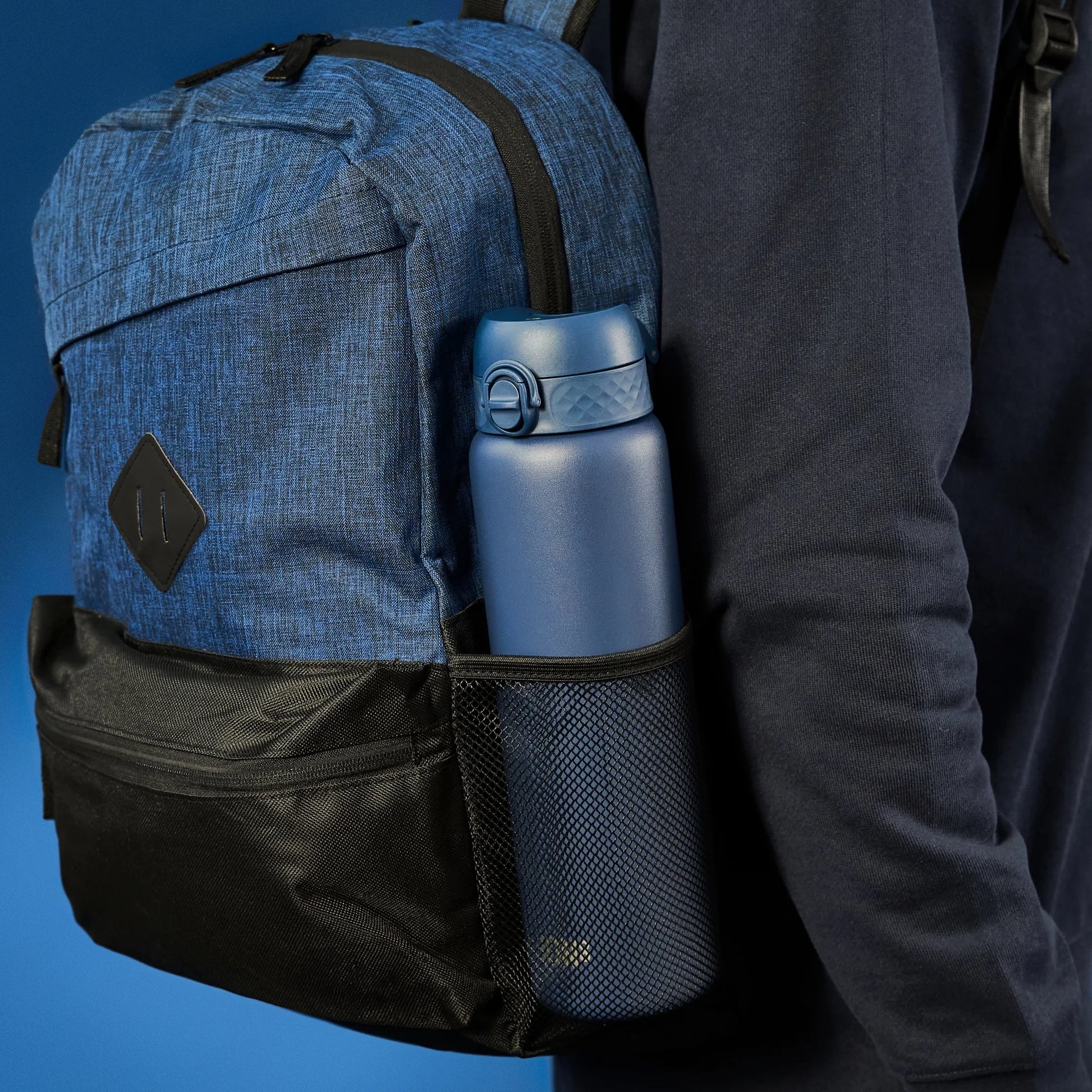 A blue backpack with a black pocket holds a blue water bottle in a mesh side pouch worn by a person in dark clothing against a blue background.