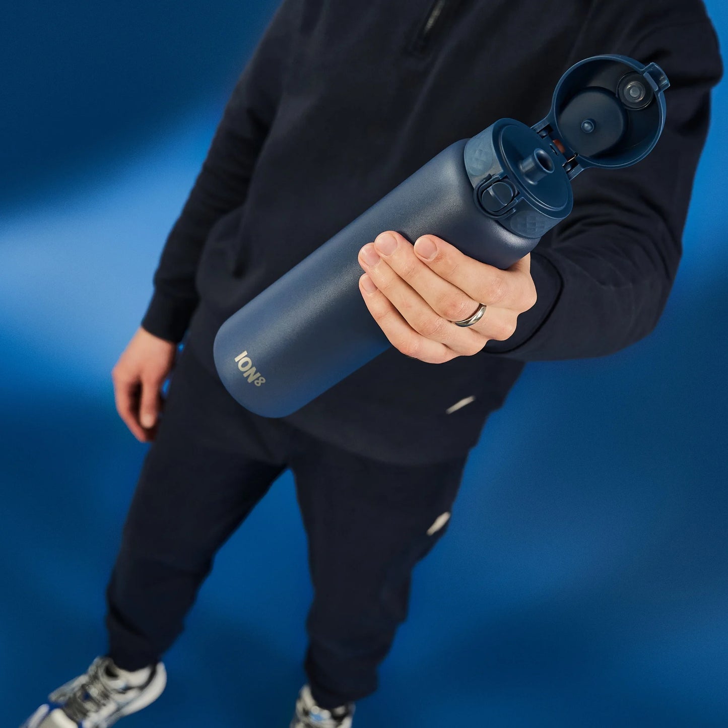 Person holding a blue water bottle with the lid open in one hand against a solid blue background wearing dark clothing. Text on the bottle reads ION8.