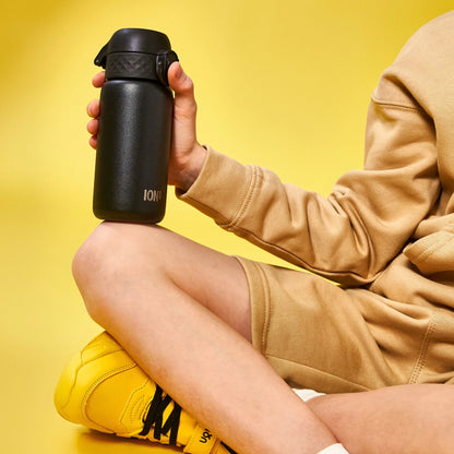 A black water bottle labeled "ION8" is held by a person in beige clothing sitting cross-legged on a yellow background wearing yellow shoes.