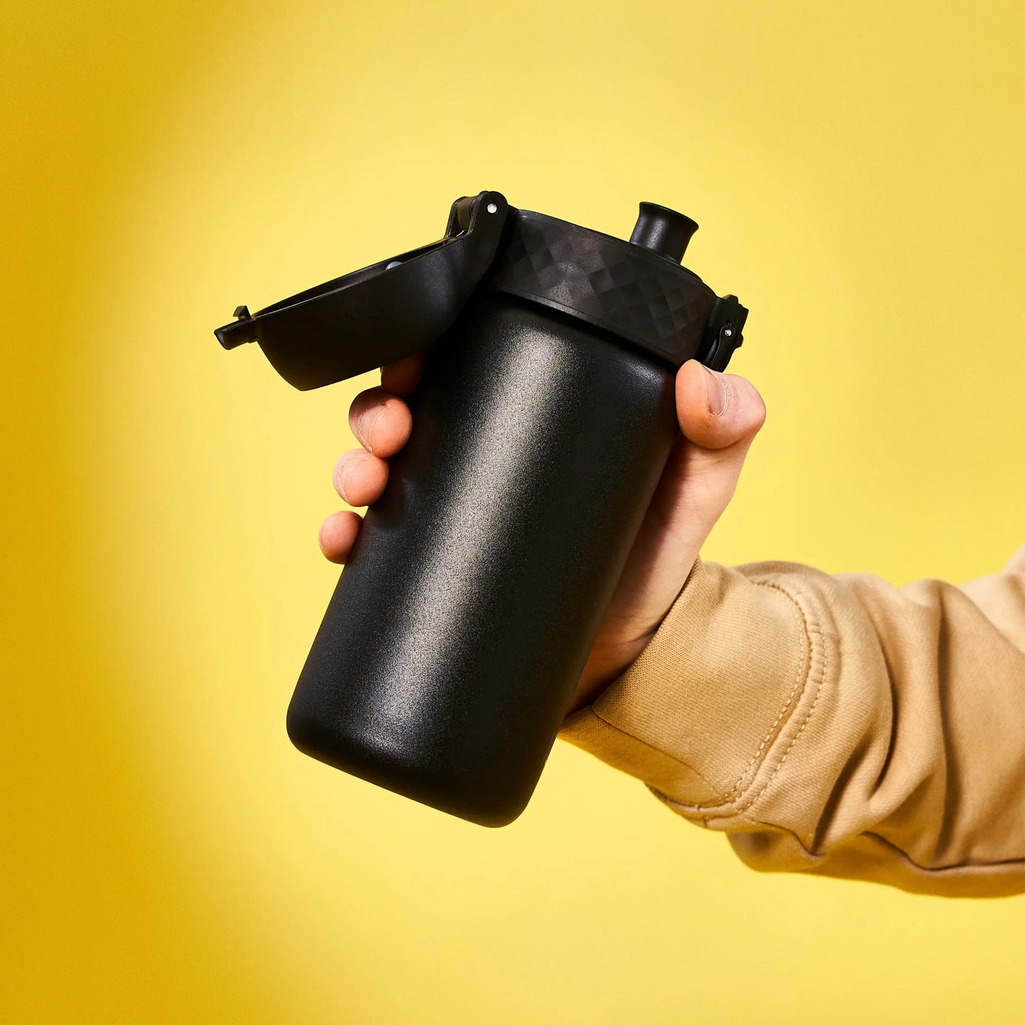 A hand holds a black water bottle with a flip-top lid against a bright yellow background