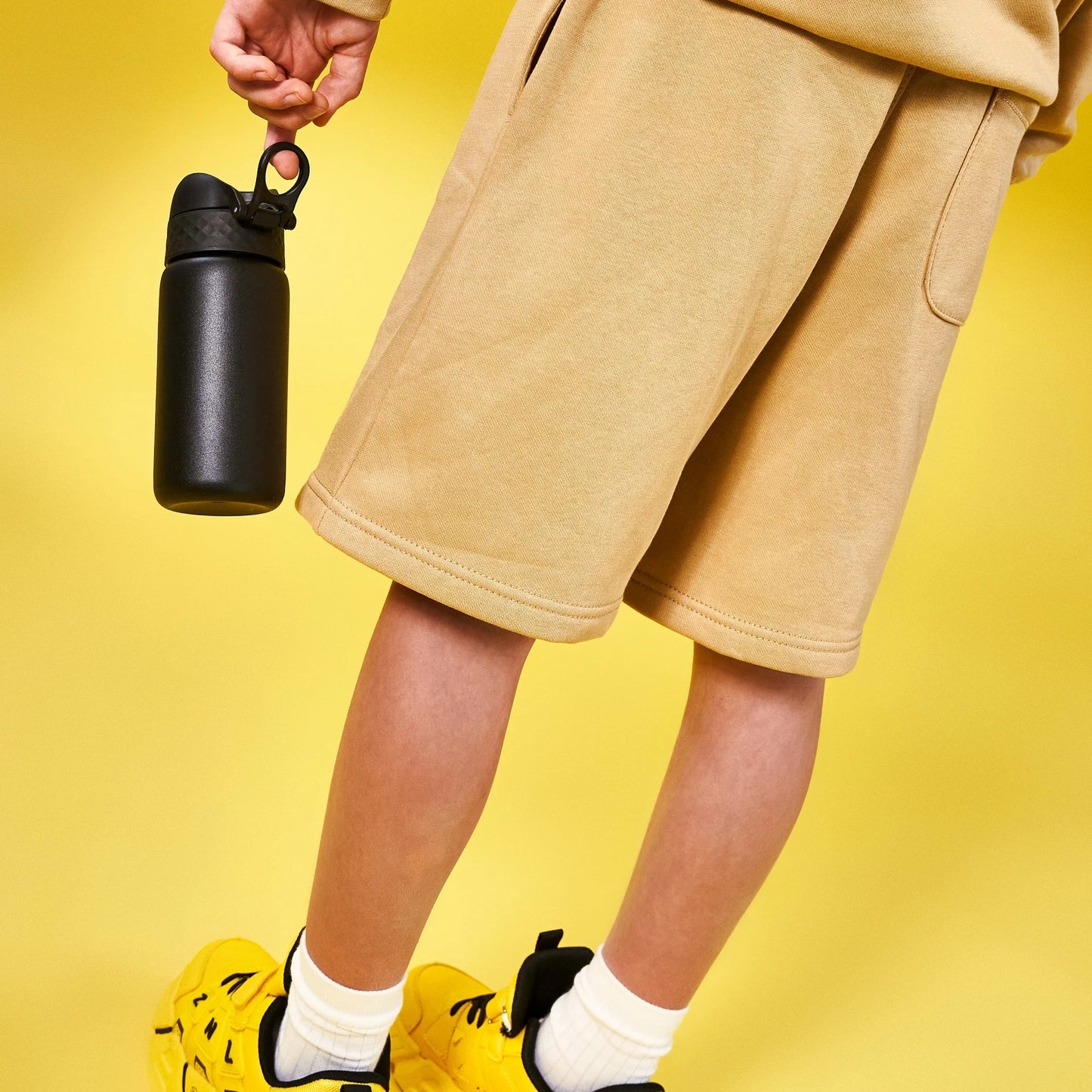 Person holds black water bottle wearing beige shorts and yellow sneakers standing against a bright yellow background.