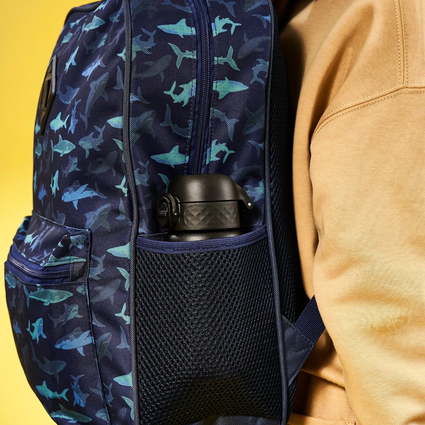 A backpack with a shark pattern holds a black water bottle in a mesh side pocket worn by a person in a tan jacket against a yellow background.