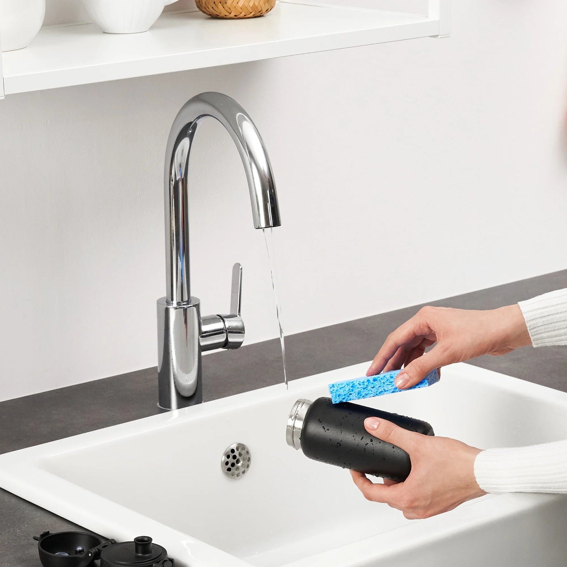 Hands are cleaning a black bottle with a blue sponge under a running kitchen faucet in a white sink with shelves above.