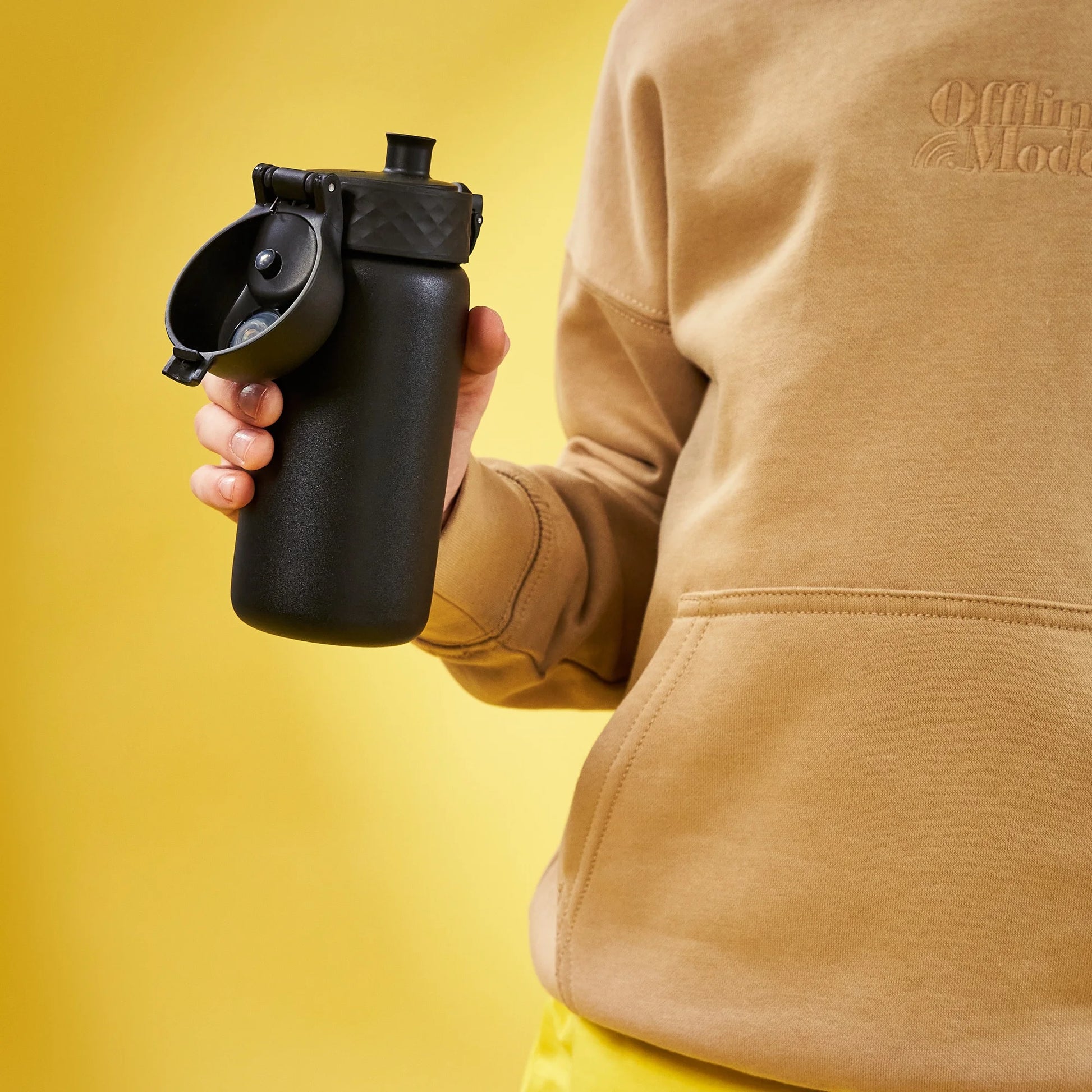 A person holds a black water bottle with a flip lid in front of a bright yellow background wearing a beige sweatshirt with the text "Offline Mode" partially visible on it.