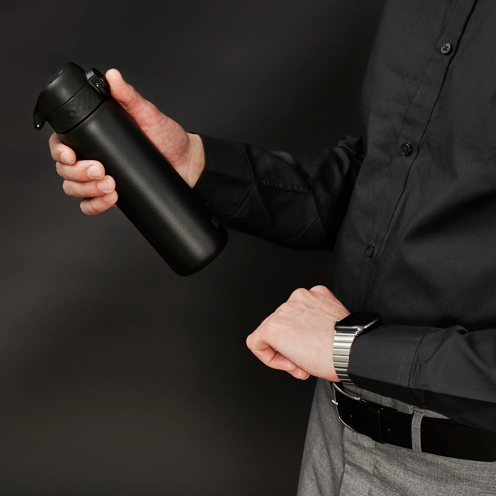 A person holds a black thermos in one hand while checking the time on a smartwatch with the other hand wearing a dark shirt and gray pants against a plain dark background