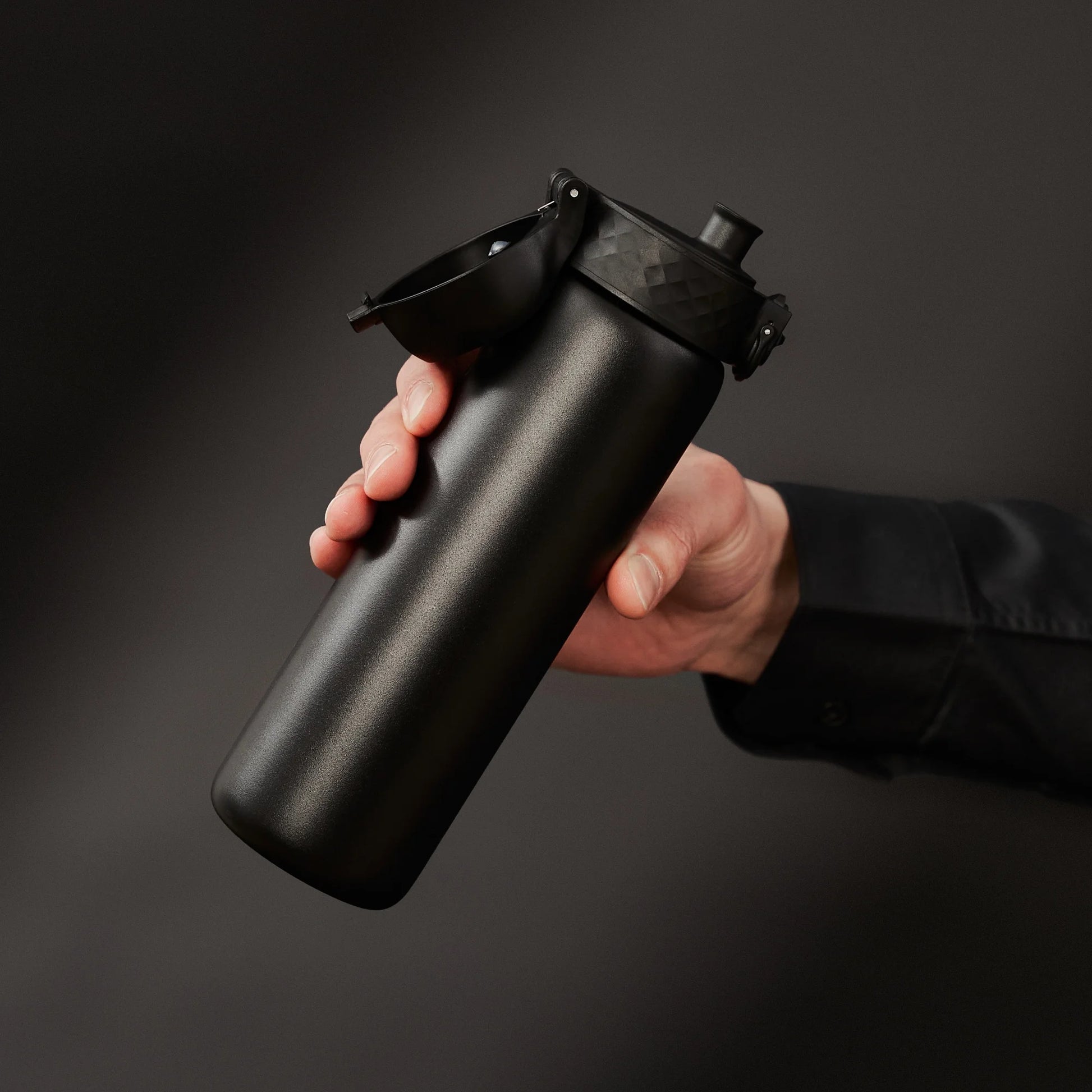 A hand holds a sleek black water bottle with a flip-top lid against a dark background, showcasing the bottle's matte finish and ergonomic design.