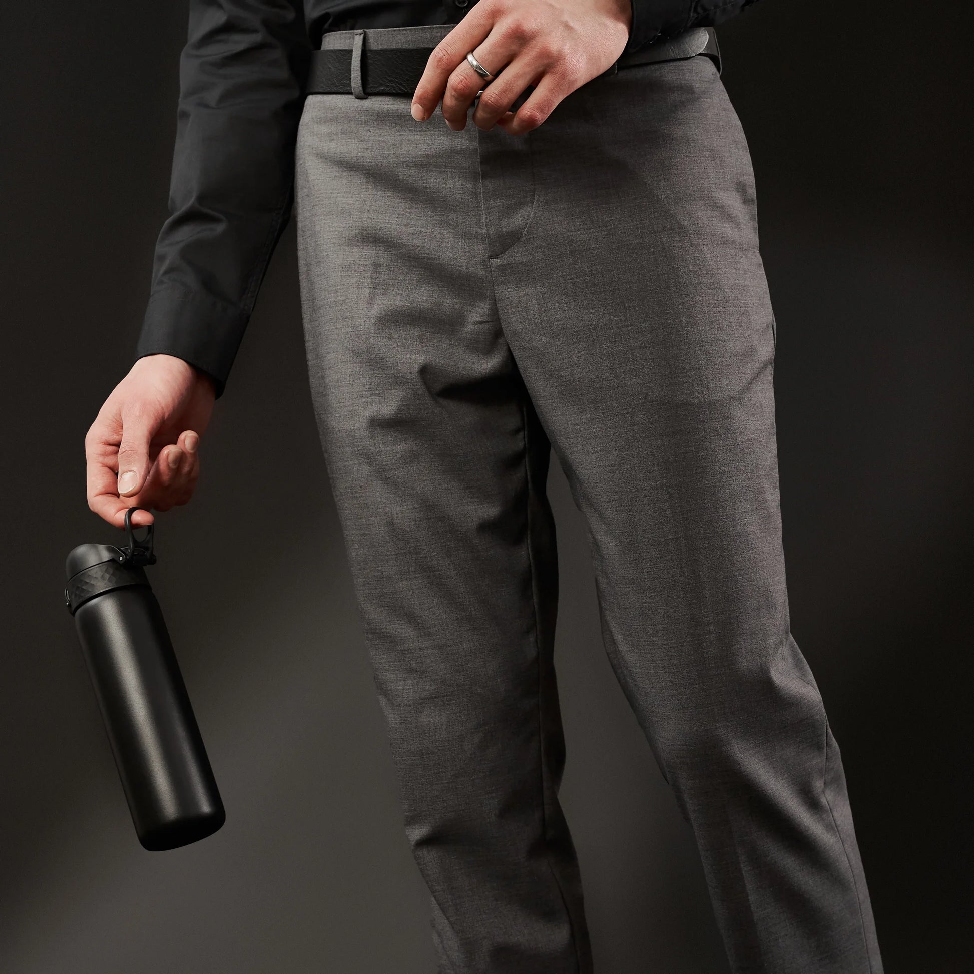 A person in gray pants and a black shirt holds a black water bottle by its loop against a dark background.