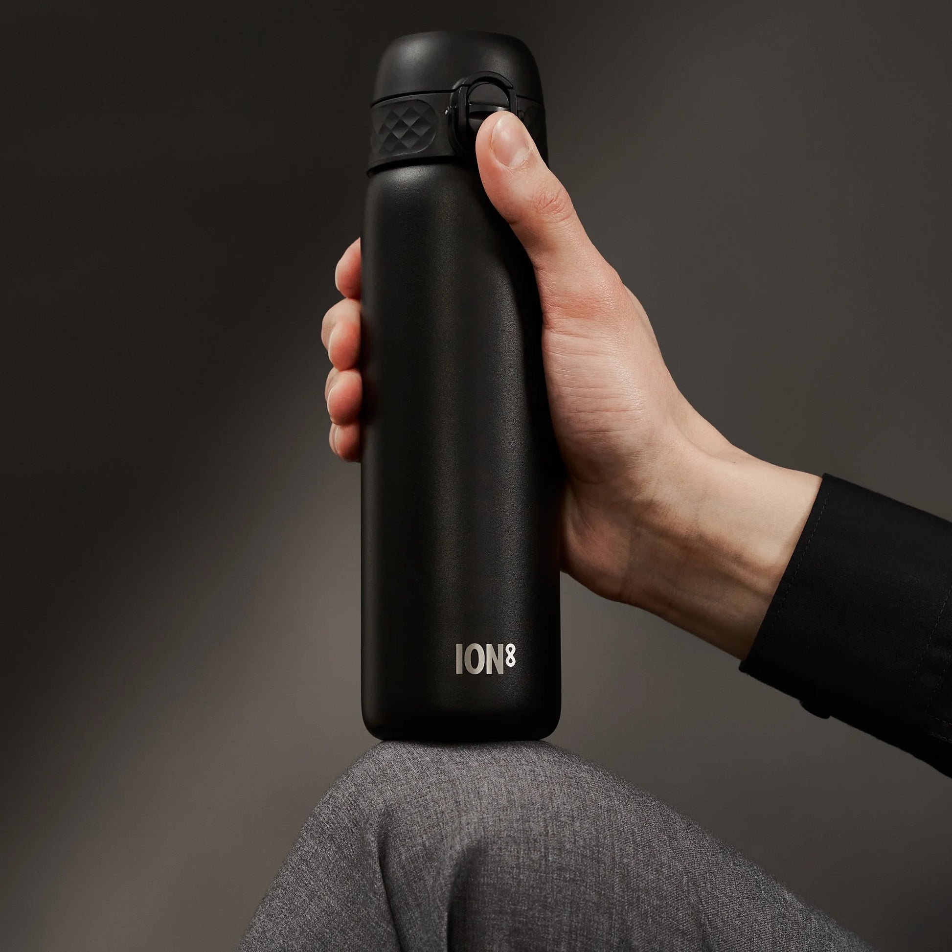 A hand holds a sleek black water bottle labeled "ION8" resting on a bent knee in a dark, neutral background.