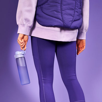 A person holds a purple water bottle by its strap while wearing a purple jacket and leggings against a purple background.