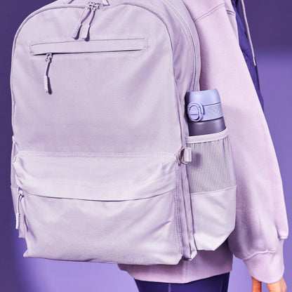 A lavender backpack hangs from a person's shoulder with a water bottle in the side pocket against a purple background.