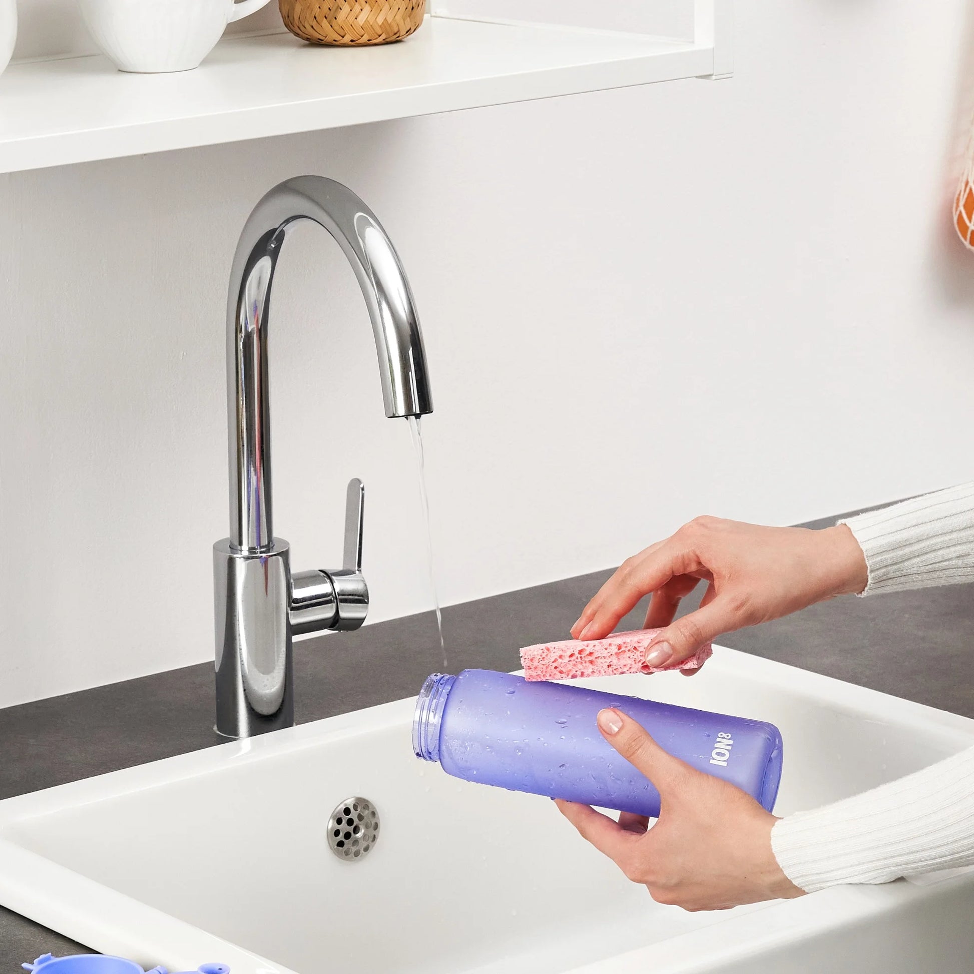 Hands scrubbing a purple water bottle with a pink sponge under running faucet in a kitchen sink setting with white shelf and countertop nearby