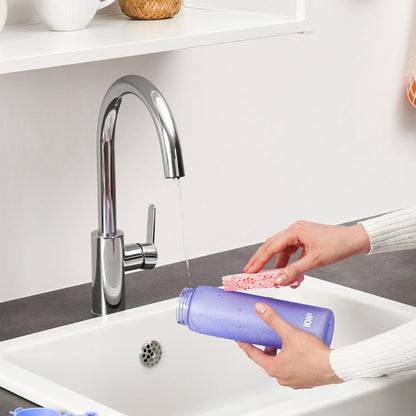 Hands scrubbing a purple water bottle with a pink sponge under running faucet in a kitchen sink setting with white shelf and countertop nearby