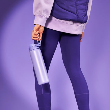 A person holds a purple water bottle by the handle wearing blue leggings and a light purple sweatshirt standing against a purple background.