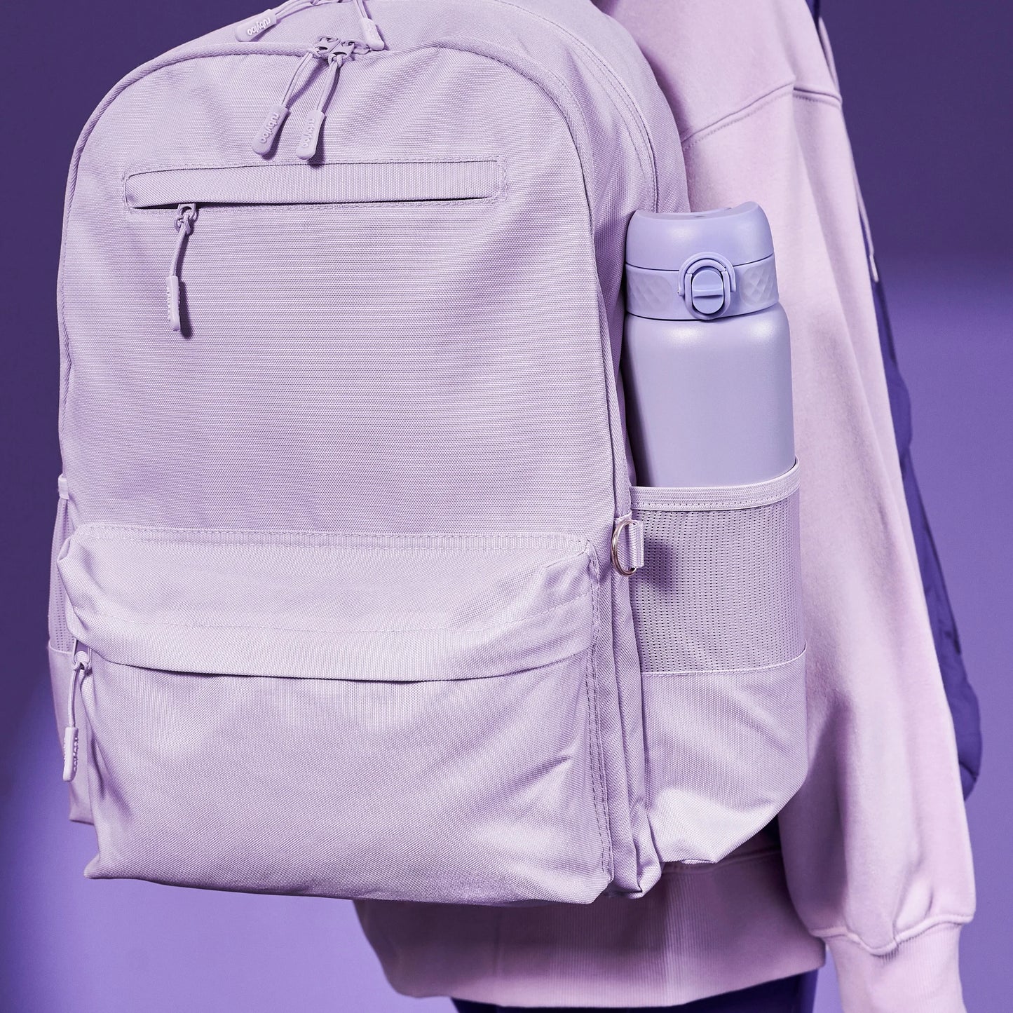 A lavender backpack hangs on a person's shoulder with a matching water bottle in the side pocket against a purple background.