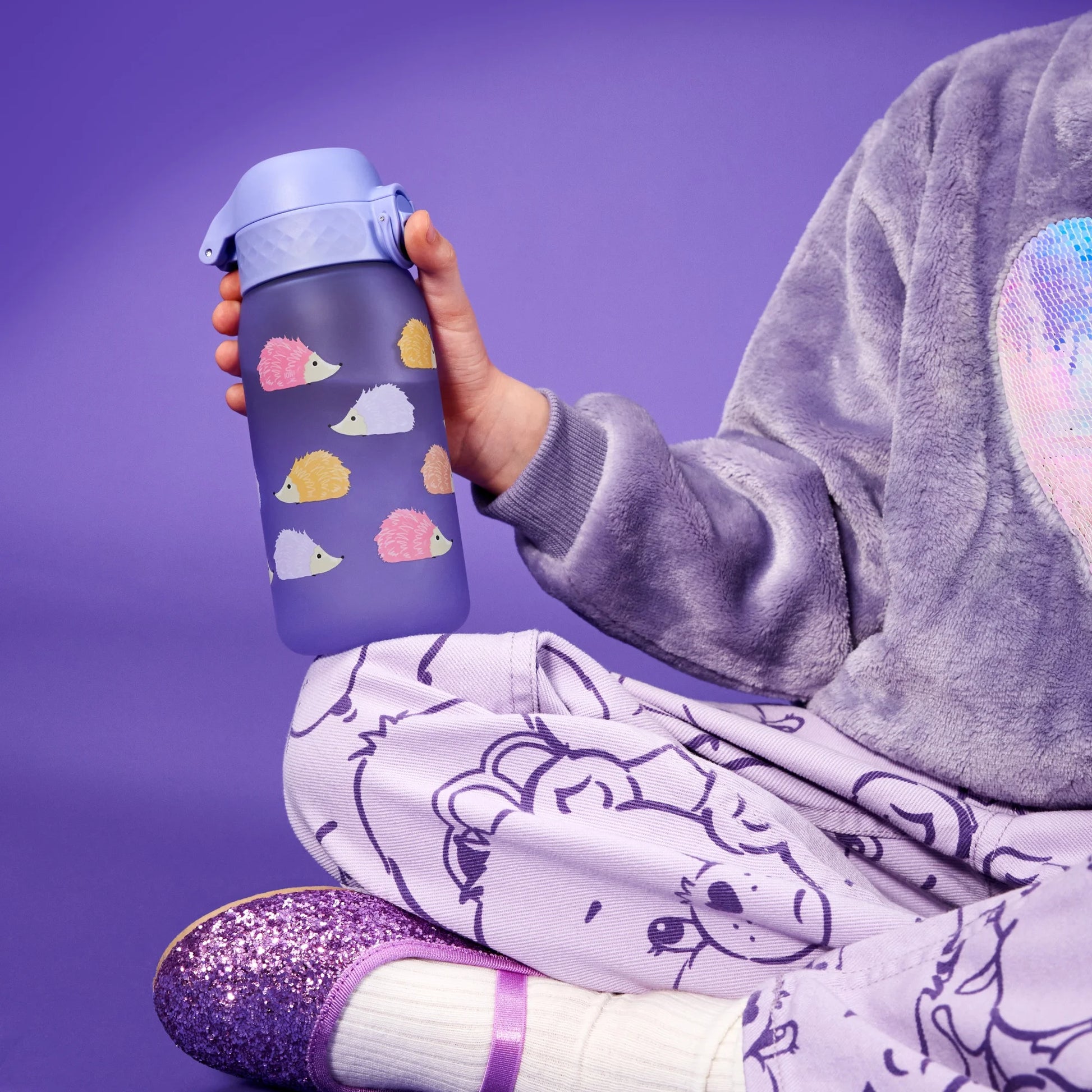 A child holds a purple water bottle with colorful hedgehog designs while sitting cross-legged wearing lavender pajamas and glittery shoes in a purple-hued setting.