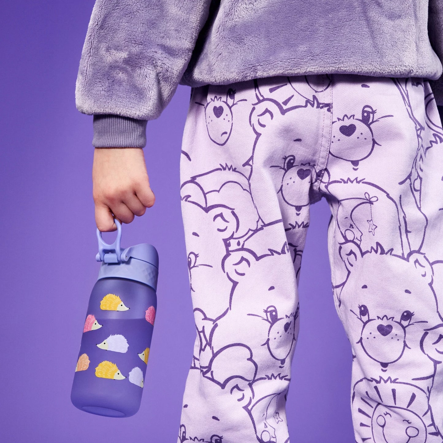 Child holds a purple water bottle decorated with colorful hedgehogs wearing a fluffy purple sweatshirt and pants patterned with cartoon bears standing against a matching purple background