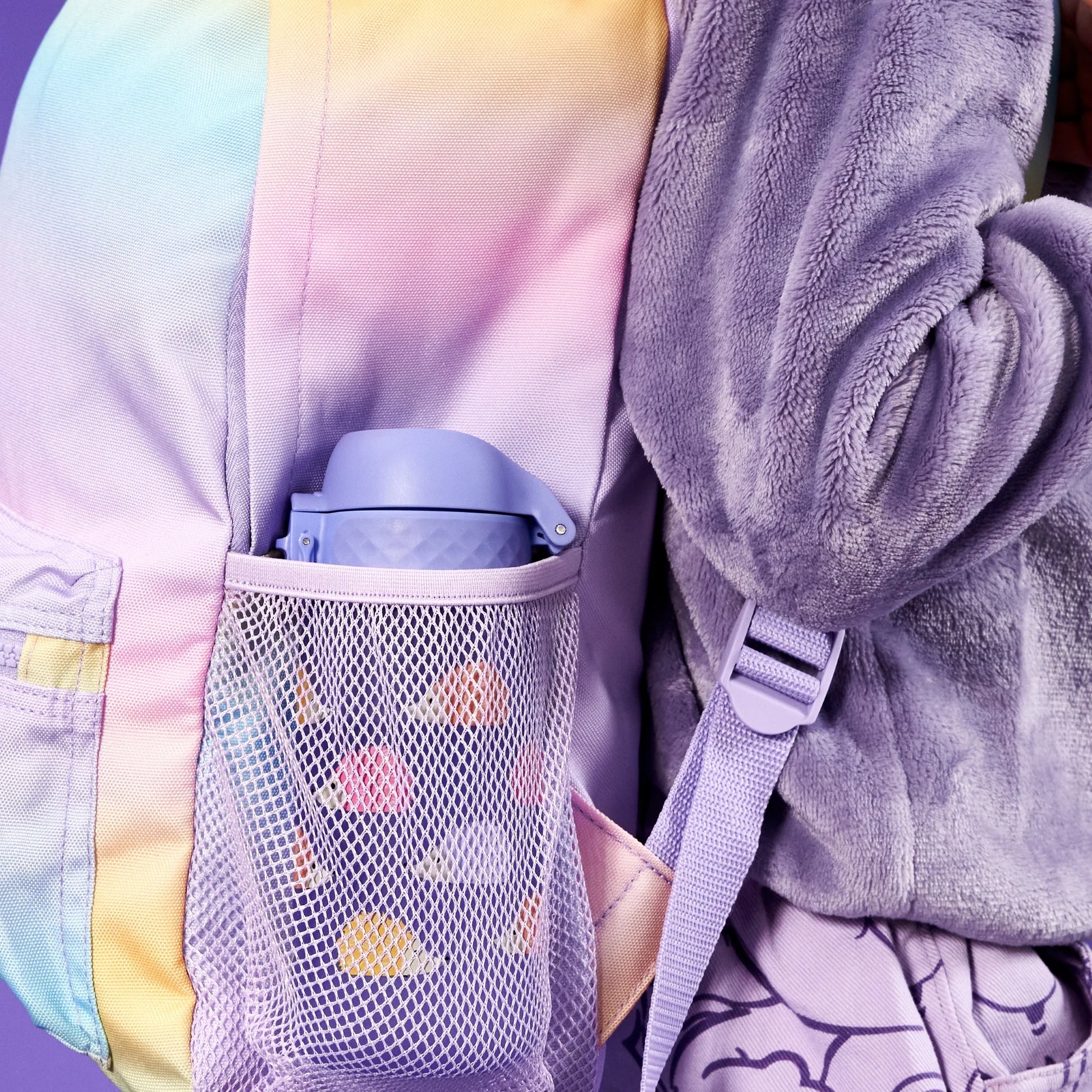 Pastel-colored backpack with mesh pocket holds purple water bottle against a purple fleece jacket worn by a person standing in a purple-themed environment.