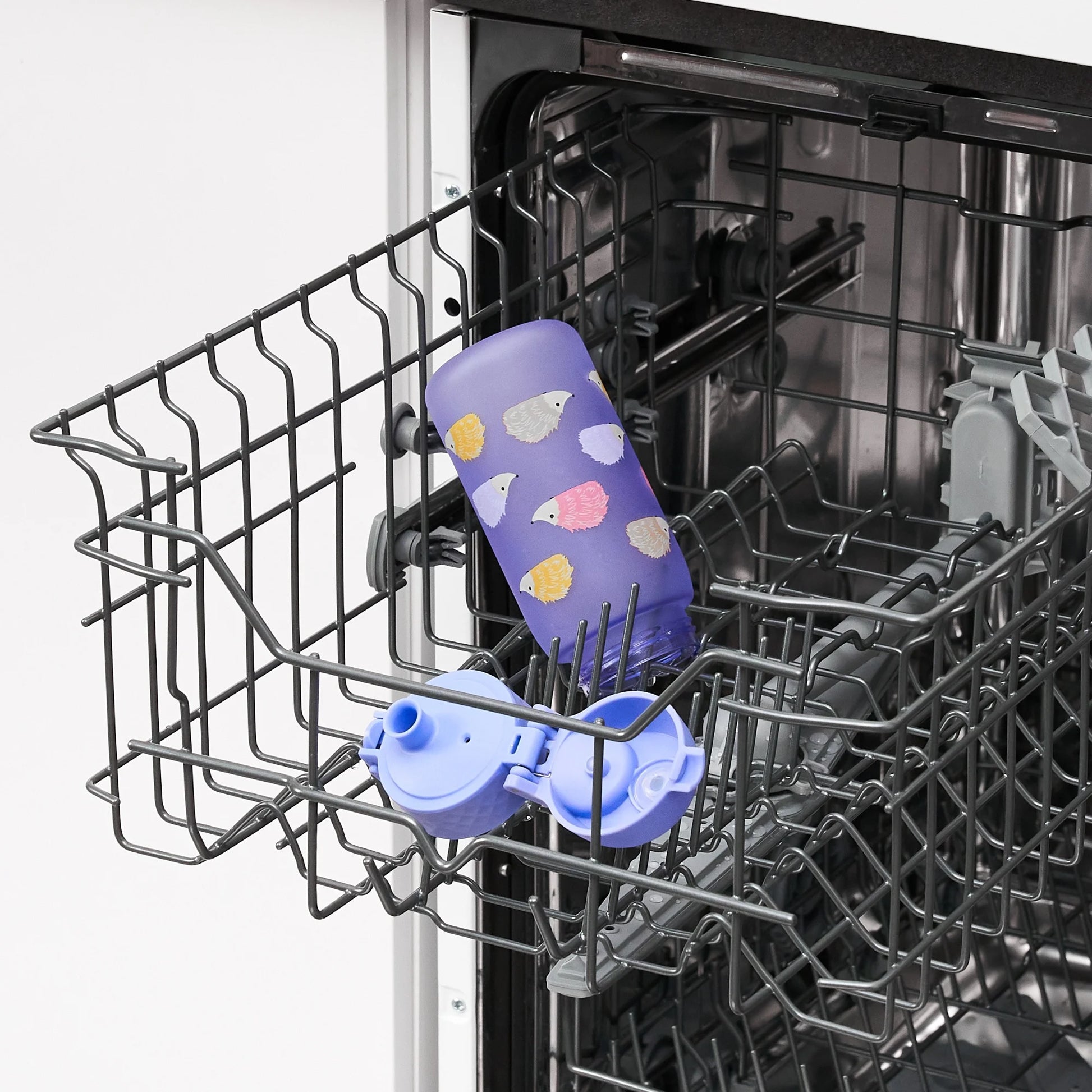 A purple bottle with illustrated sheep lies open on the top rack of a dishwasher, alongside its separated cap, in a kitchen setting.