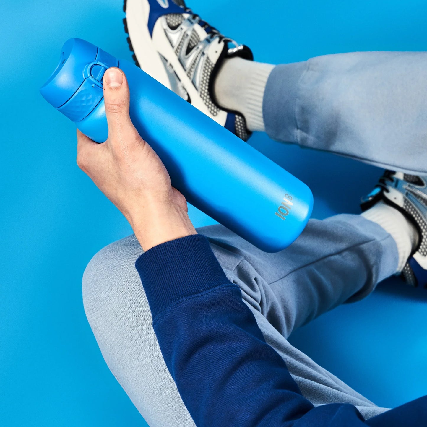 A blue water bottle is held by a person wearing a blue sweater and light pants sitting on a blue floor. Sneakers and the number 1018 are visible.