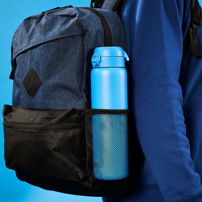 A blue water bottle sits in a mesh side pocket of a dark blue backpack worn by someone in a blue hoodie against a plain blue background.