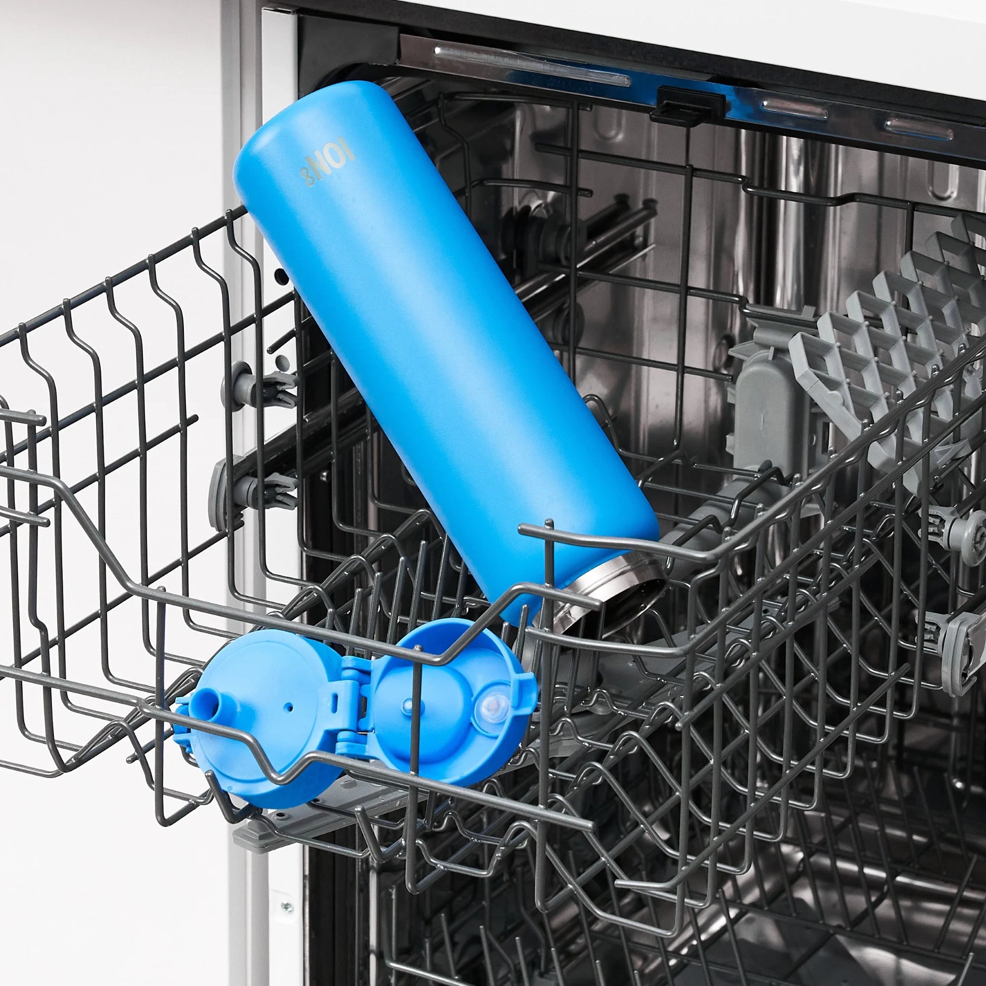 A blue water bottle lies inside an open dishwasher rack, surrounded by dish racks and a detachable bottle lid. Text on the bottle reads 301.