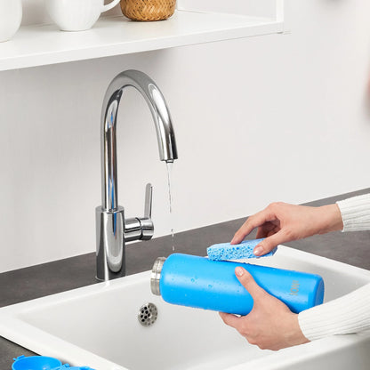 Hands holding a blue bottle with a sponge cleaning it over a white sink under a flowing chrome faucet in a kitchen setting