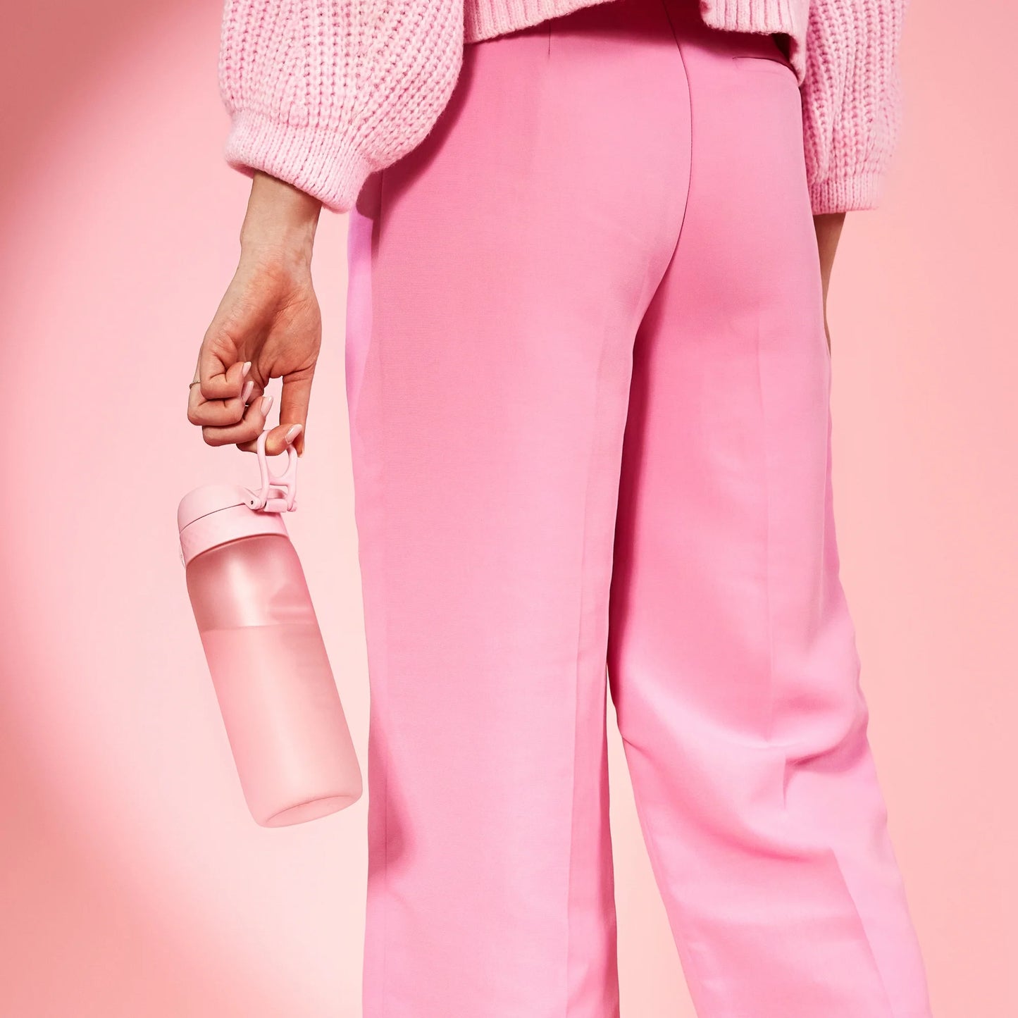 A pink water bottle is held by a person wearing pink pants and a knitted sweater against a pink background.