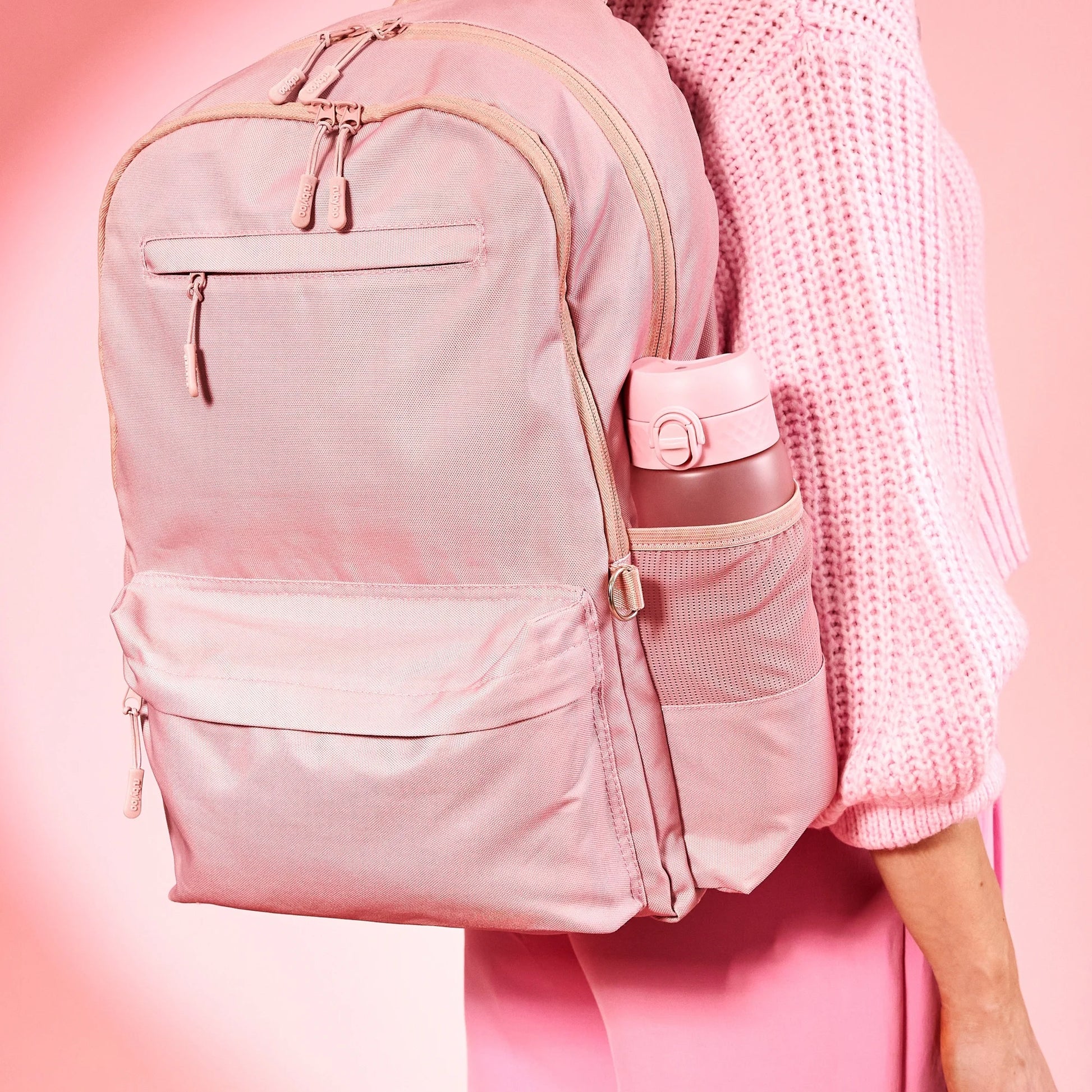 A person carries a large pink backpack with a water bottle stored in its side pocket wearing a pink sweater and pants standing against a pink background.