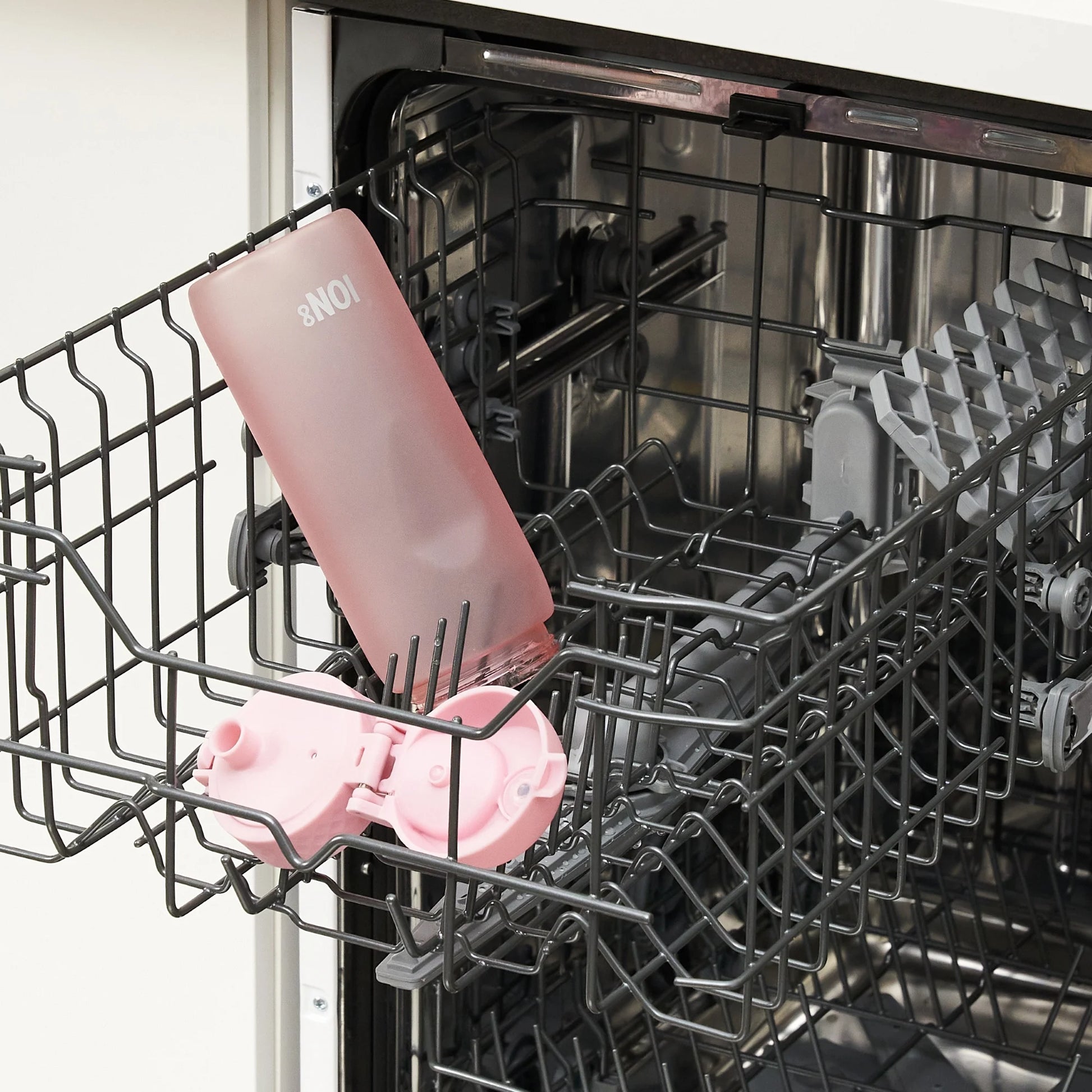 A pink water bottle marked "8NOI" is positioned inside an open dishwasher, resting on the top rack among gray dish supports in a kitchen setting.