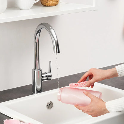 Hands scrub a pink bottle with a sponge under running water in a white kitchen sink with a modern chrome faucet nearby shelves holding mugs and a basket