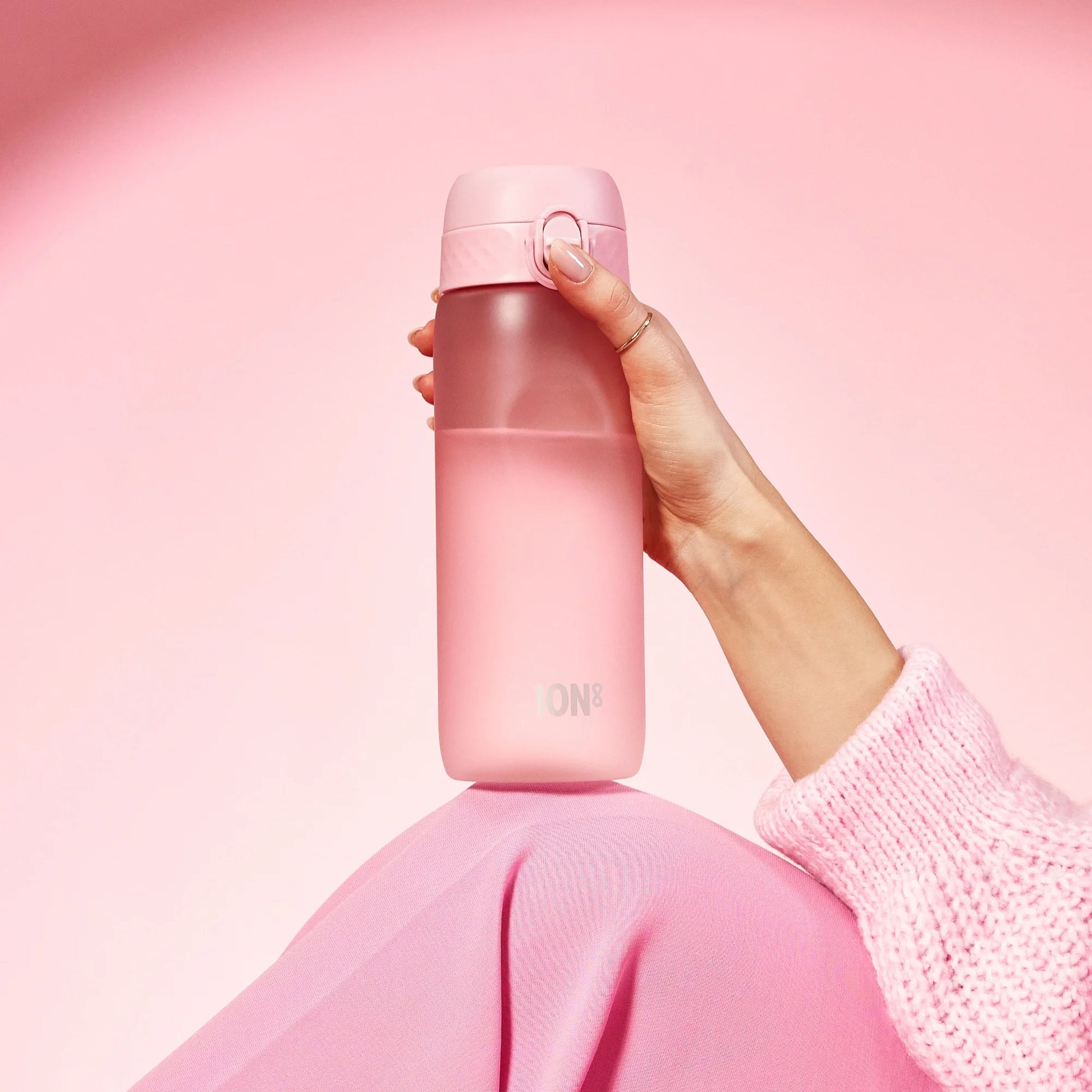 Hand holding a pink water bottle marked with "ION8" against a pink background while wearing a pink sweater draped over a knee.