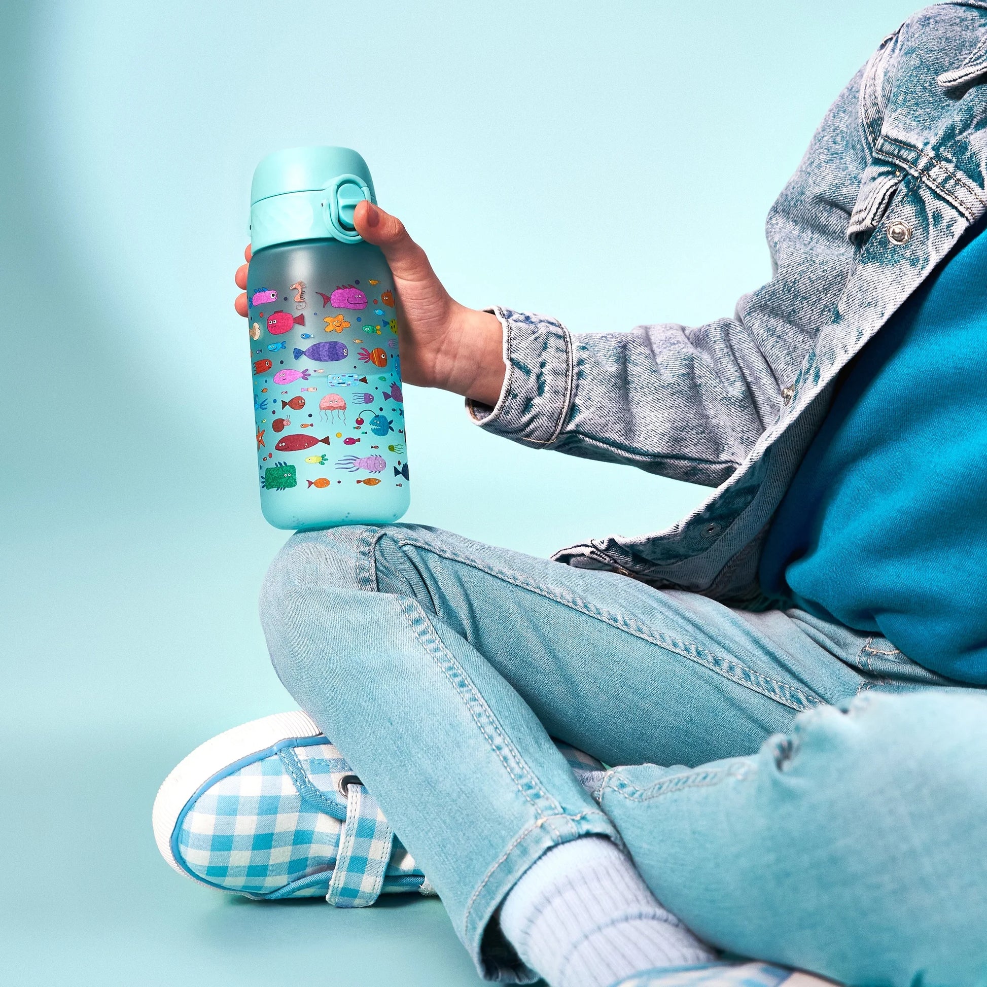 A colorful water bottle featuring sea creatures held by a person wearing light denim and seated cross-legged on a light blue background.