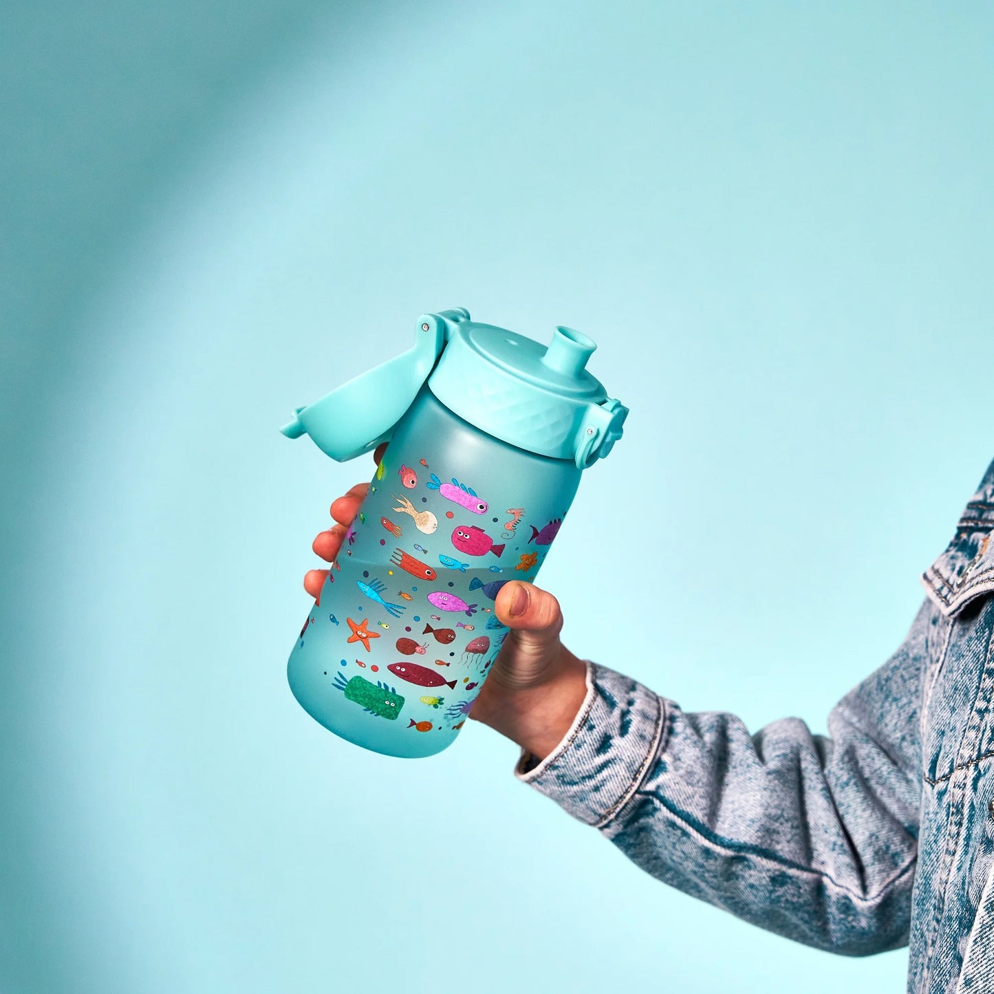 A blue water bottle with colorful cartoon sea creatures held by a person wearing a denim jacket against a matching blue background.