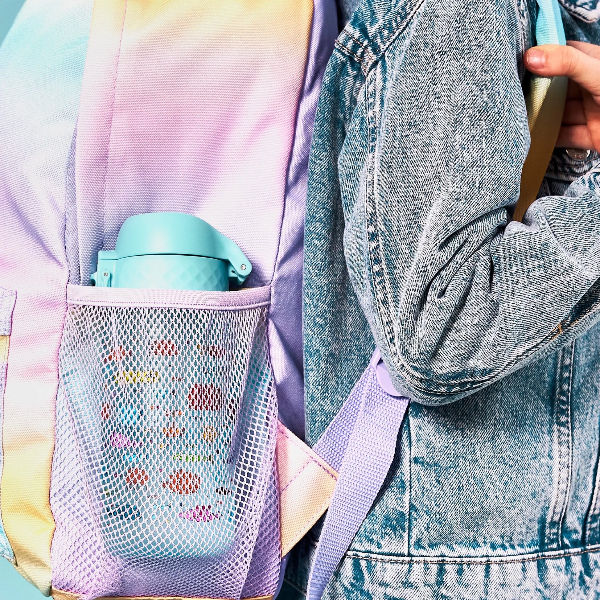 A multicolored backpack holds a light blue water bottle in its mesh side pocket, next to a person wearing a denim jacket who is adjusting a shoulder strap.