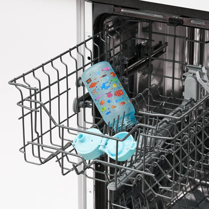 A colorful baby bottle lies open inside the top rack of a dishwasher in a kitchen setting.