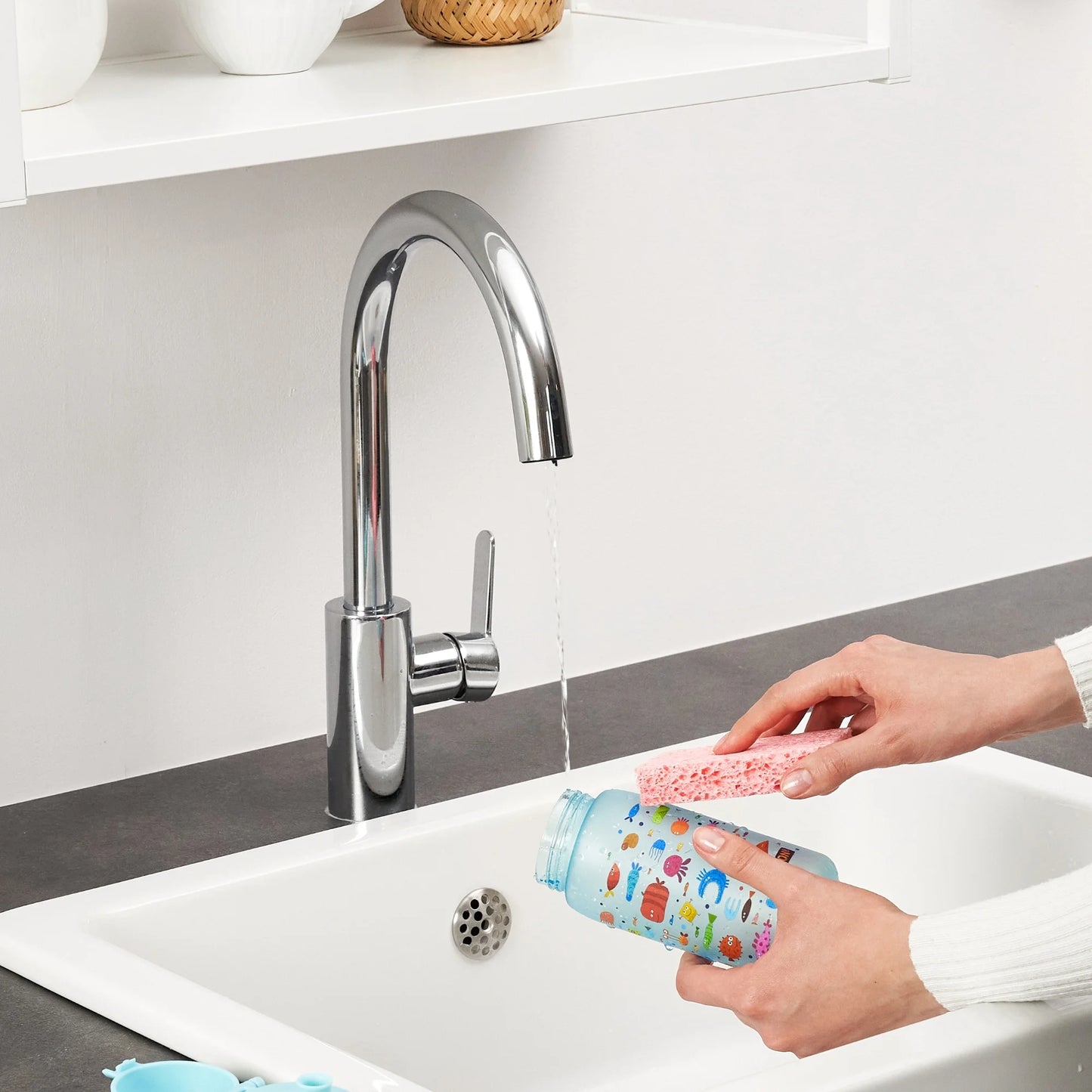 Hands cleaning a colorful baby bottle with a sponge under a running faucet in a modern kitchen sink setting