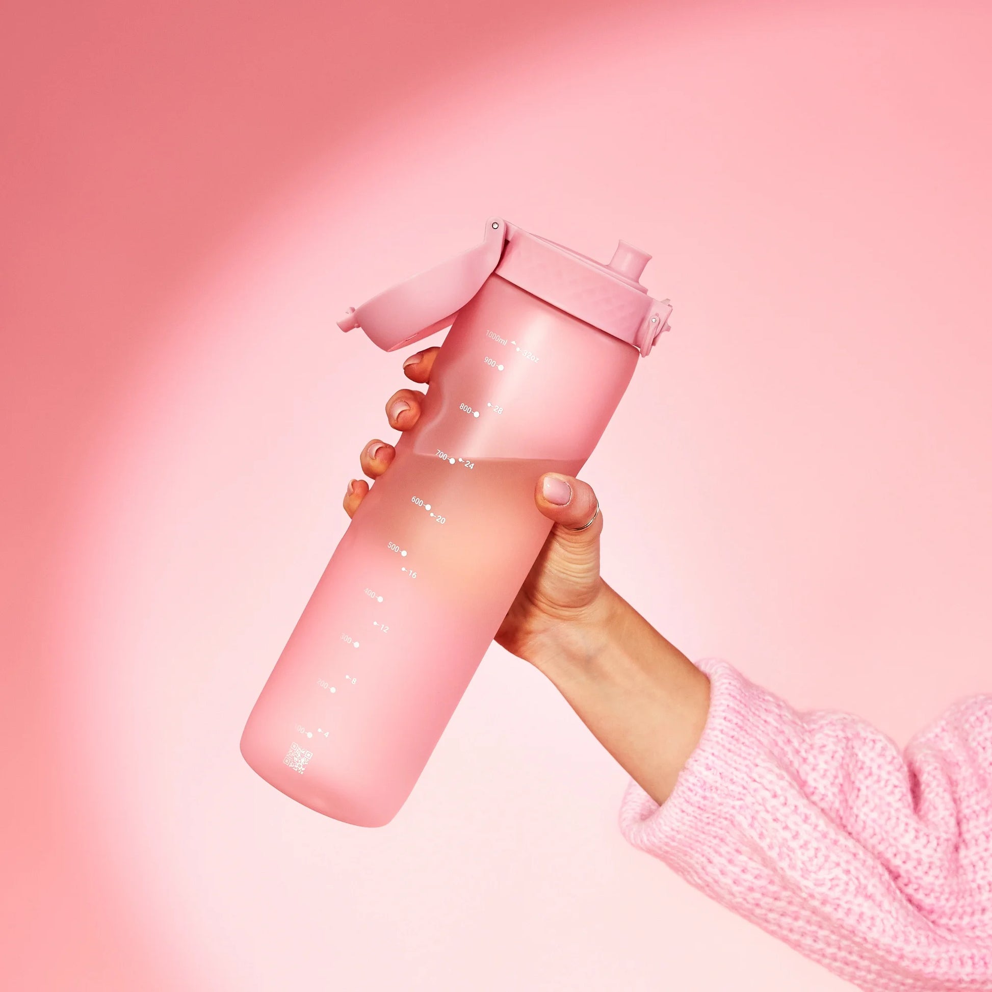 A pink water bottle with time markers is held by a hand wearing a pink sweater against a soft pink background