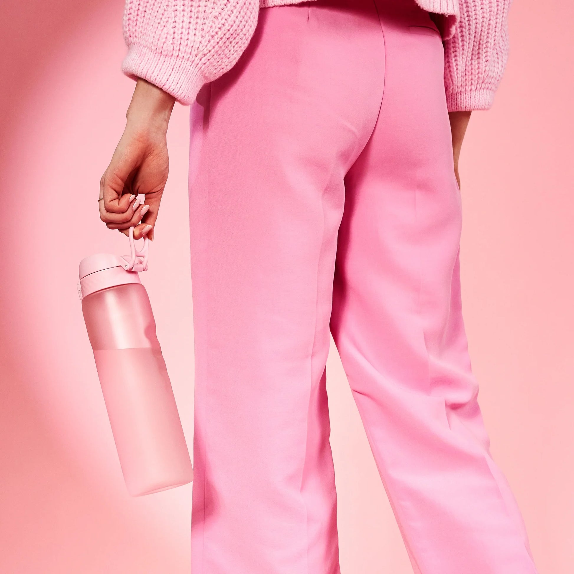 A person holds a pink water bottle by its loop in a hand, wearing pink pants and a pink sweater, set against a pink background.