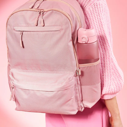 A pink backpack worn by a person in a pink sweater and pants features a side pocket holding a matching pink water bottle set against a pink background.