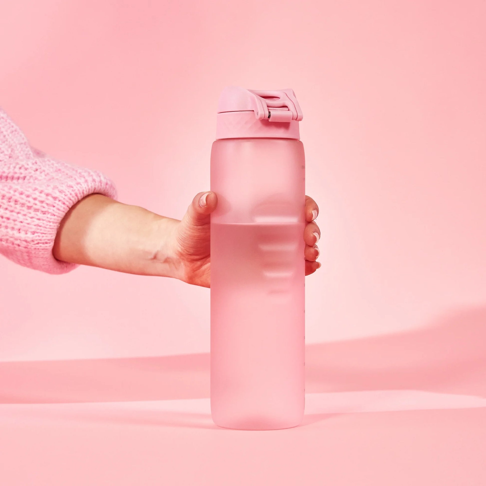 A hand holds a pink water bottle with a flip-top lid against a solid pink background wearing a pink knit sweater.